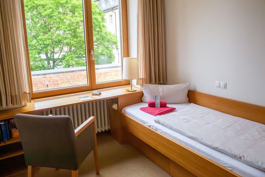 Blick auf ein Bett, daneben steht ein Sessel und darüber befindet sich ein Fenster mit Blick auf einen Baum
